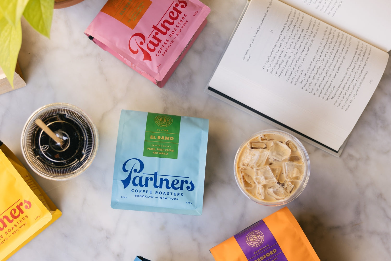 Partners Coffee Our Favorite Resources blog post. This image displays bags of Partners Coffee retail bags paired with a cold brew and iced latte ordered by a customer reading a book.